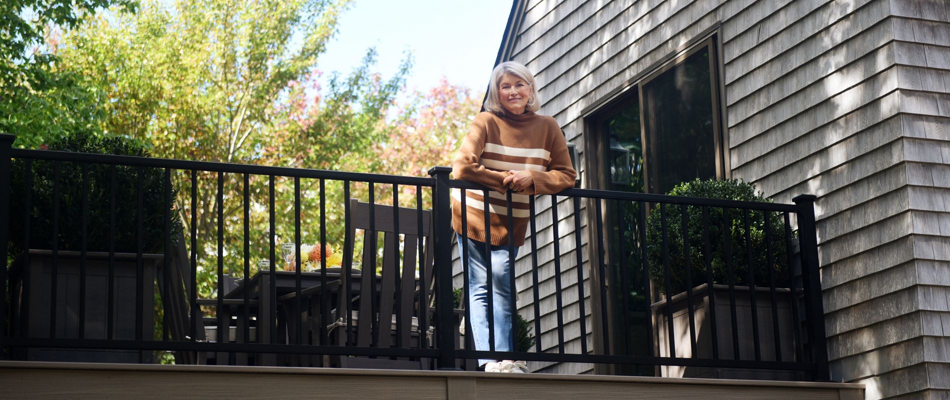An older woman stands on a raised balcony attached to a house with wooden shingle siding. She leans casually on the black railing, smiling toward the camera. Potted plants and outdoor furniture decorate the balcony, suggesting a comfortable outdoor living space. Sunlight filters through surrounding trees, creating a bright, relaxed atmosphere.